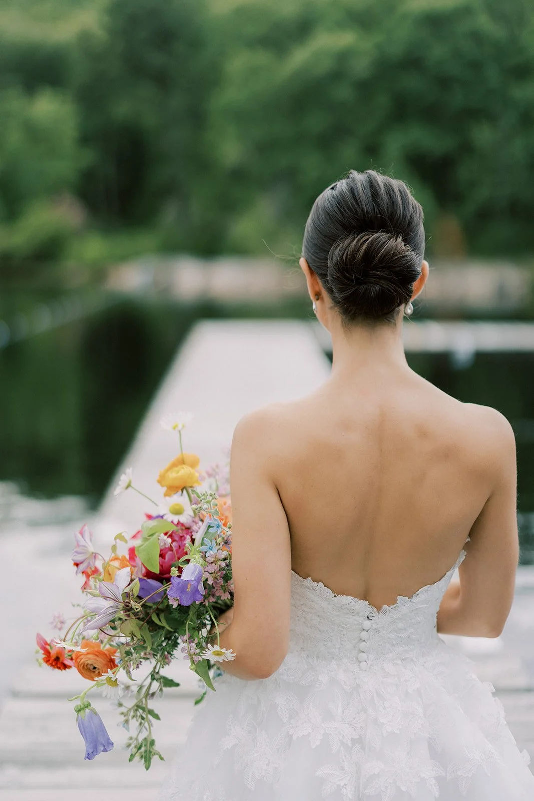 Back view of a bride holding a colorful bouquet, wearing a strapless white wedding dress with embroidered lace, standing outdoors near water with greenery in the background.