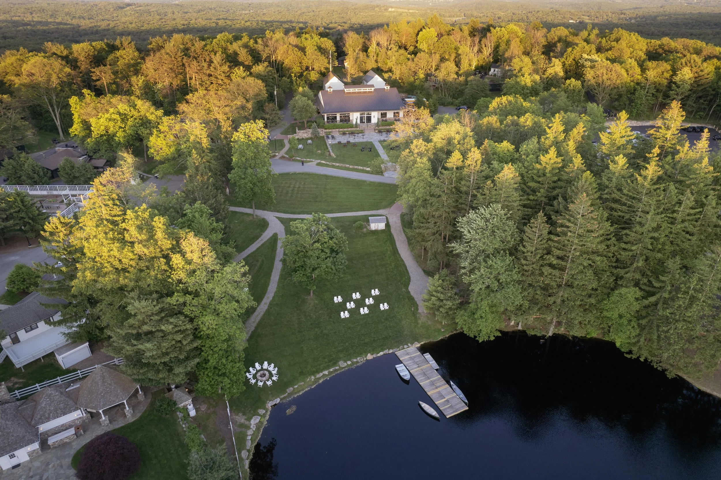 Aerial view of a lakeside area with a large house, trees, gardens, pathways, and outdoor seating in a lush, green landscape.