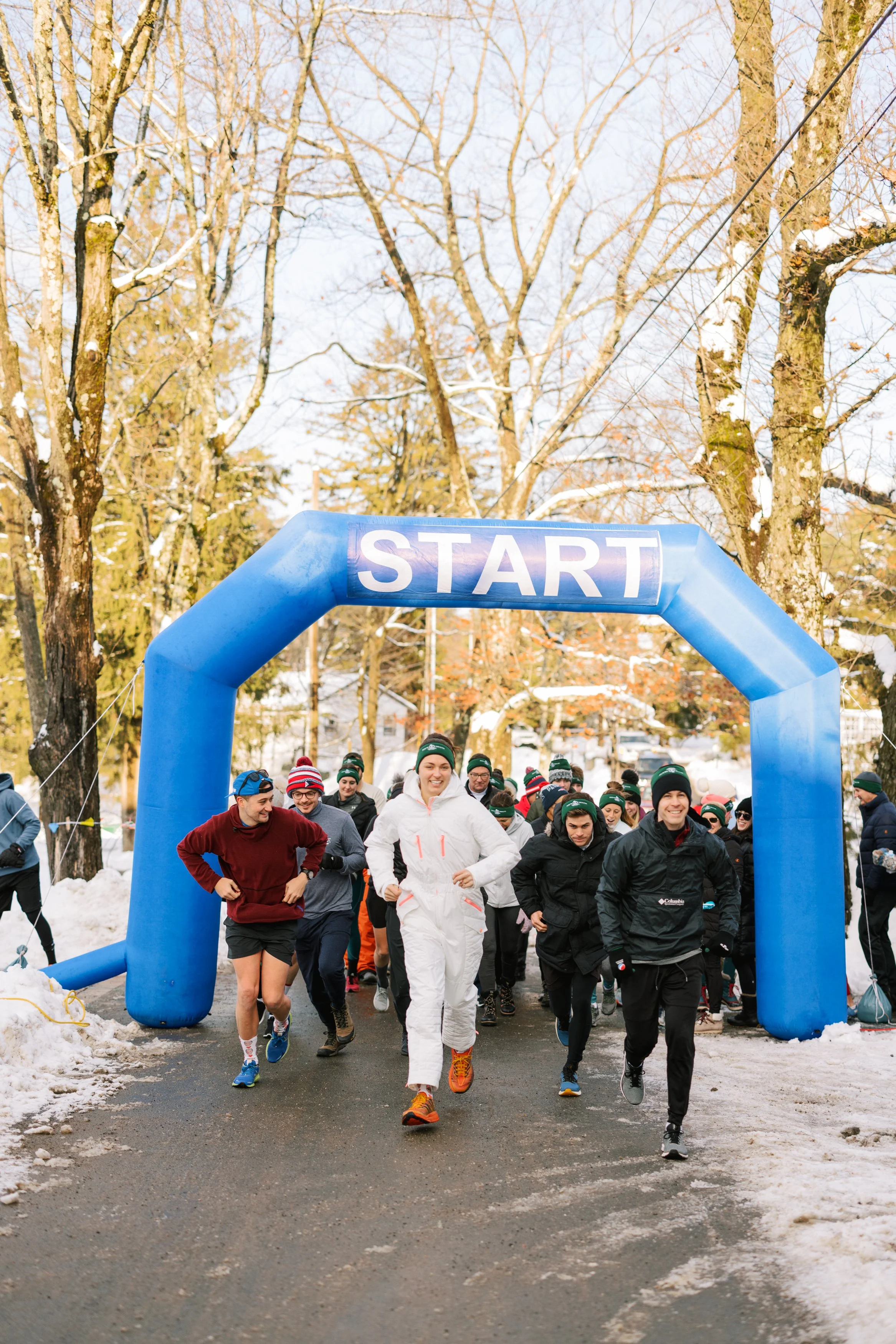 A group of runners starting a race under a blue inflatable arch labeled 'START' on a snowy outdoors path surrounded by leafless trees.