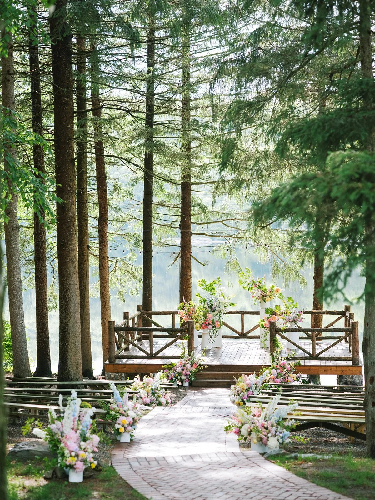 An outdoor wedding ceremony setup in a forest with a wooden platform decorated with pink and white flowers, surrounded by trees and benches.