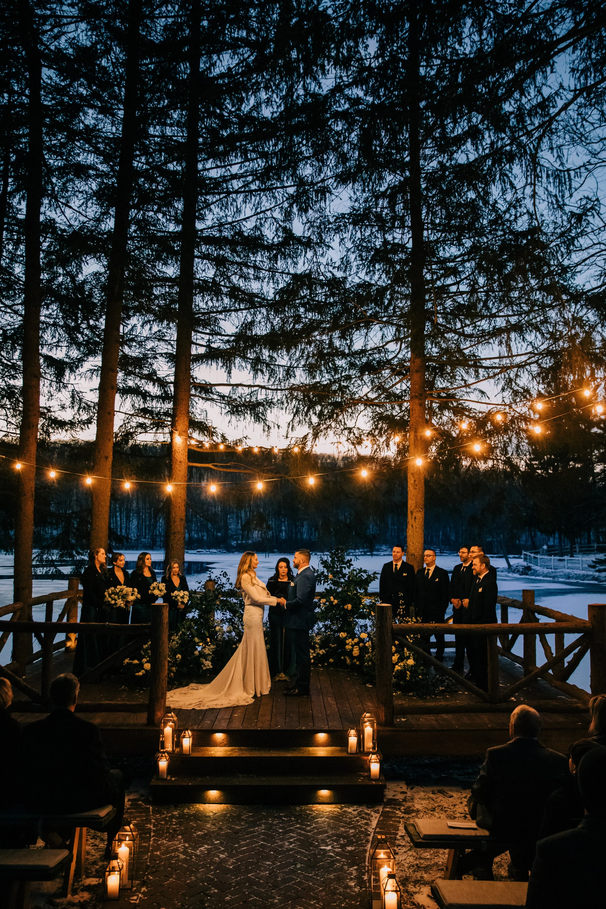 A wedding ceremony taking place outdoors at twilight, with a bride and groom holding hands in front of an officiant, surrounded by bridesmaids and groomsmen, under string lights among tall trees, with guests seated nearby and candles lining the aisle