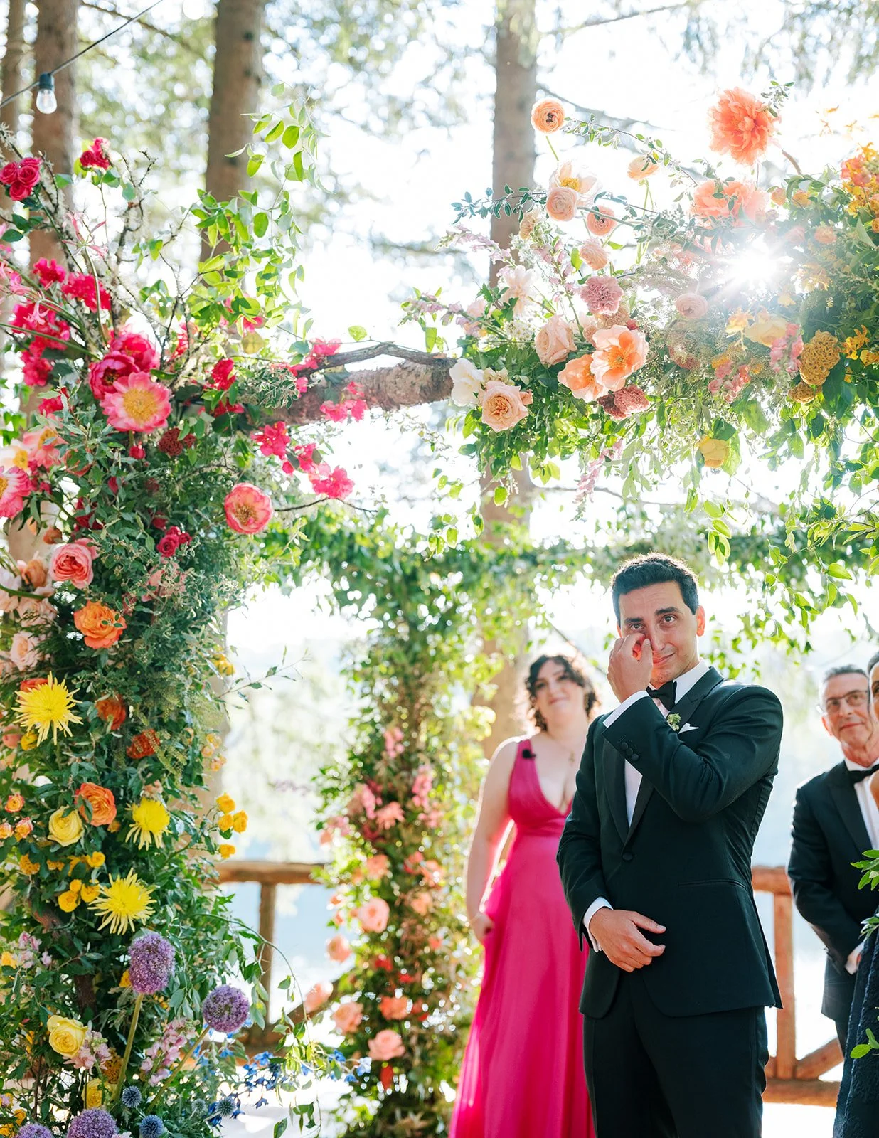 A groom dressed in a black tuxedo, wiping away tears and smiling in an outdoor wedding ceremony surrounded by colorful flowers and greenery, with bridesmaids and groomsmen in the background.