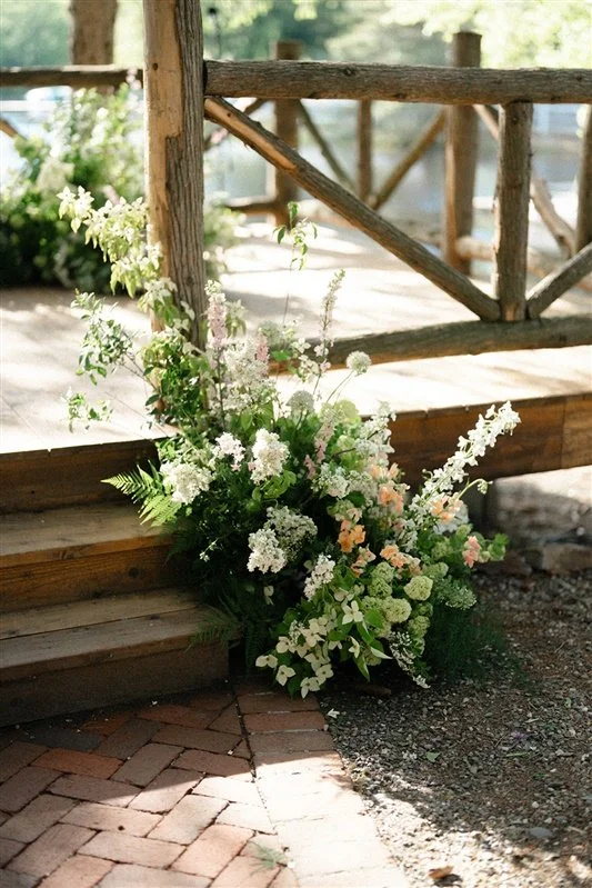 A large floral arrangement with white and pale pink flowers and green foliage, placed at the base of wooden steps next to a rustic wooden railing.