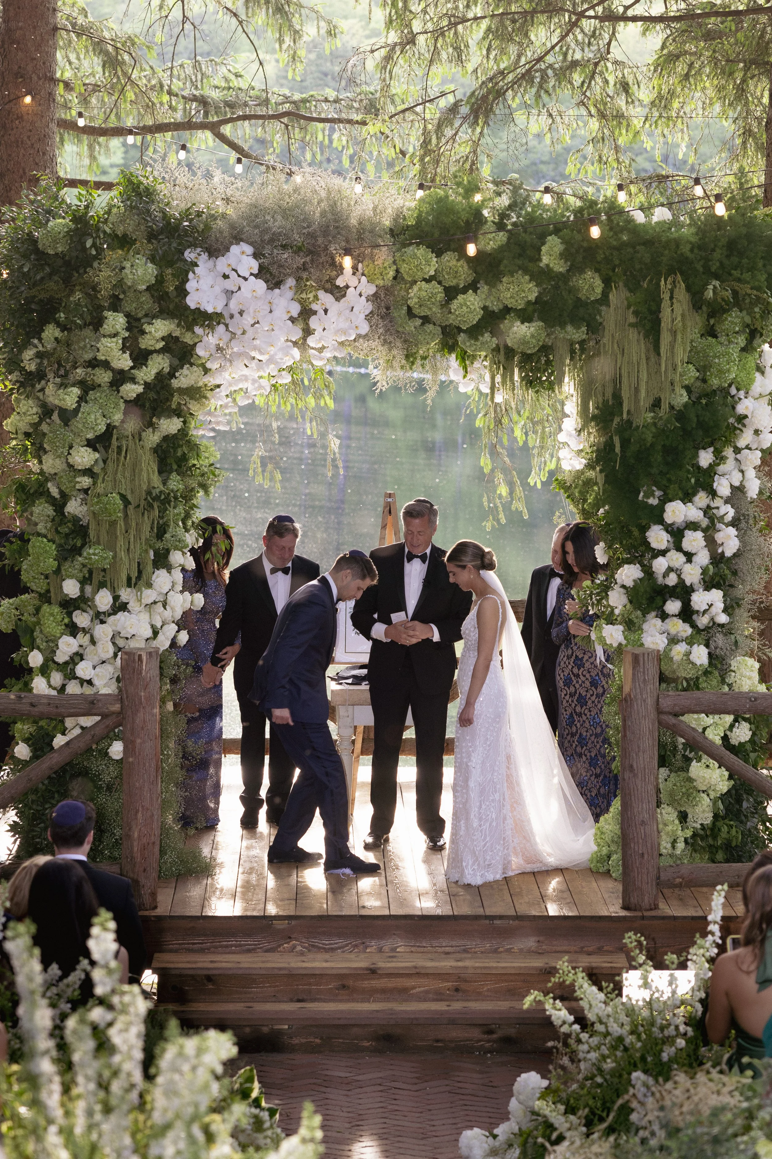 A wedding ceremony takes place outdoors on a wooden platform decorated with lush white and green floral arrangements and hanging greenery, set by a scenic lake with trees. The bride and groom, dressed in traditional wedding attire, are exchanging vow
