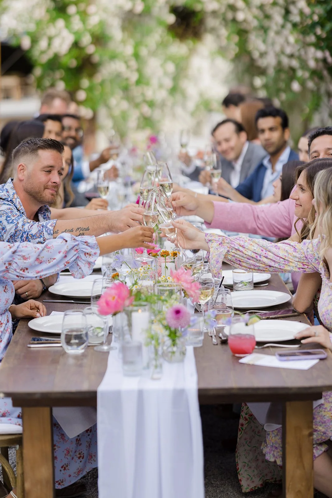 People gathered around a long outdoor table, raising glasses in a toast during a celebration, with floral centerpieces and greenery overhead.