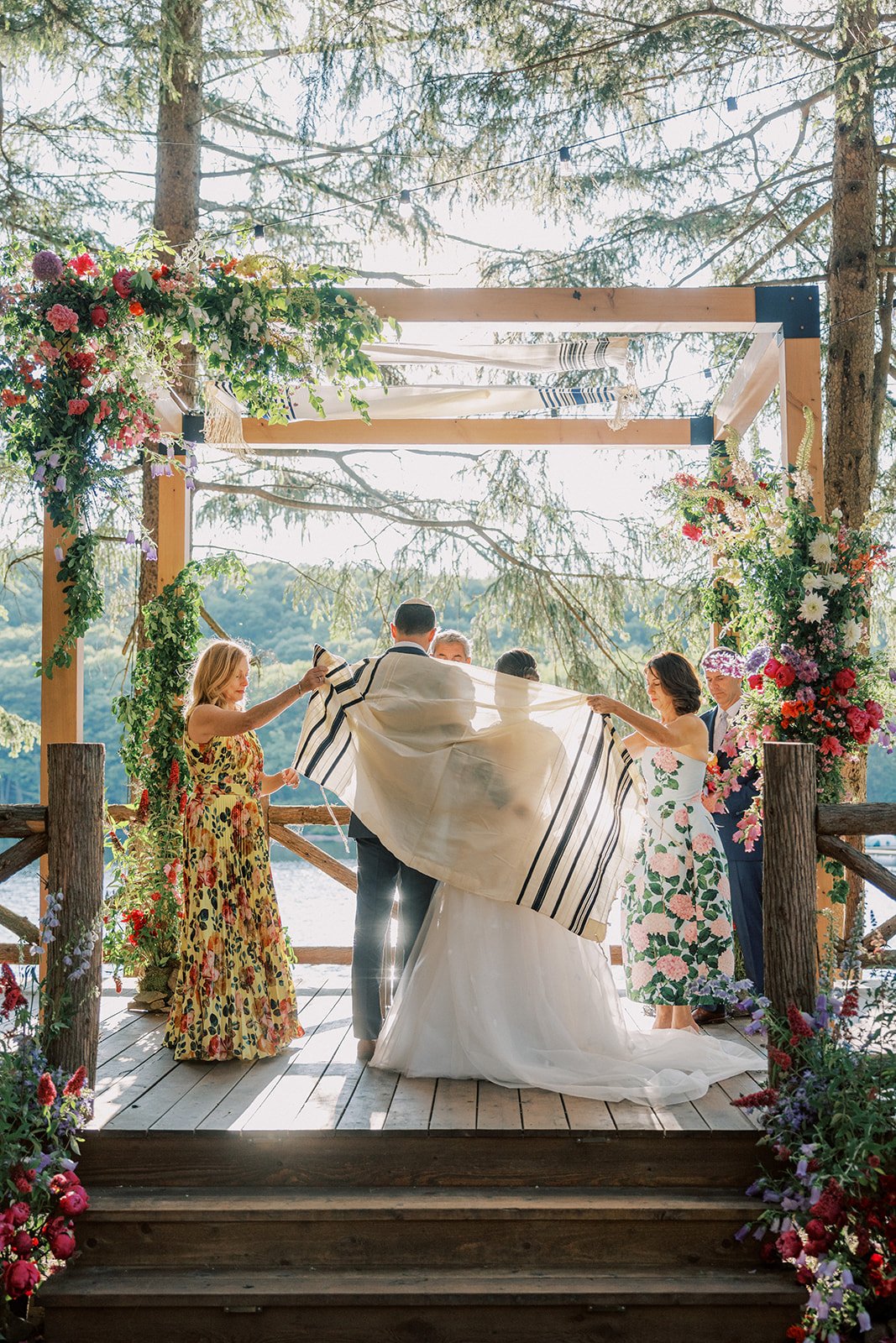 A bride and groom are standing under an outdoor wedding arch surrounded by friends and family, as the bride holds up her veil. The setting is by a body of water with trees and floral decorations.