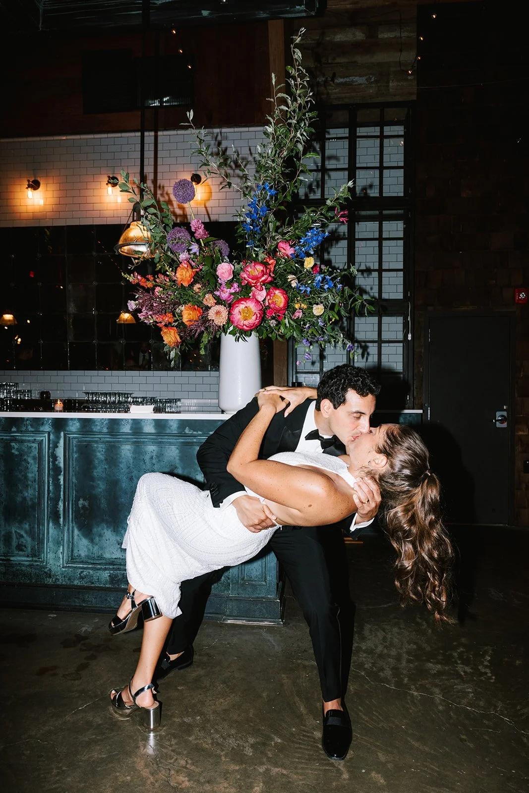 A man in a tuxedo dips a woman in a white dress for a dance in front of a large floral arrangement in a dimly lit venue.