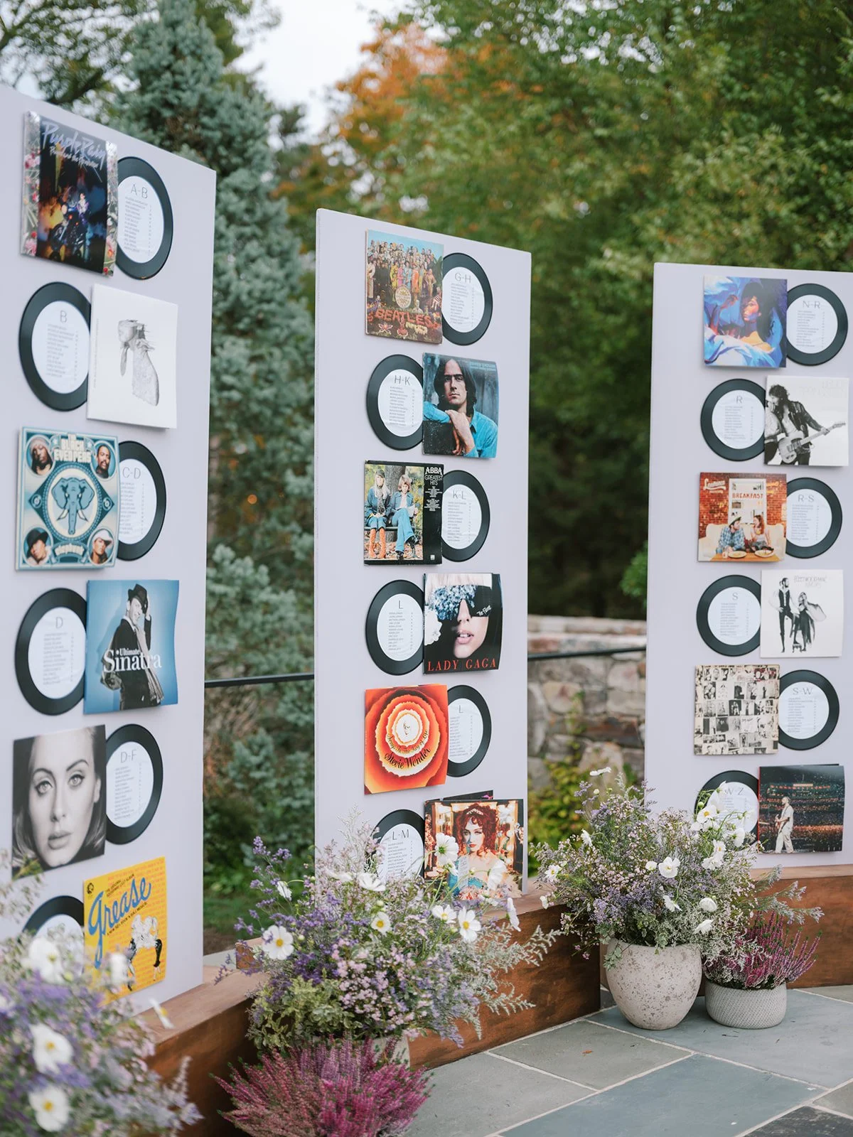 Outdoor display of vinyl records and album covers on white panels, with flower arrangements at the base, set against trees with fall foliage.