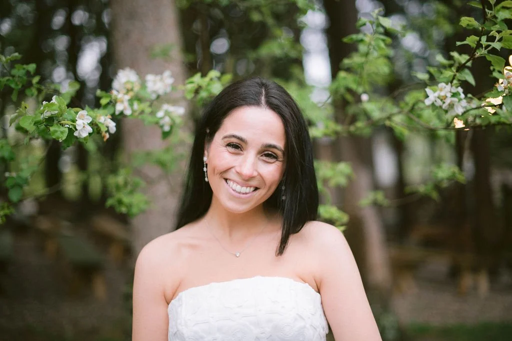 A woman with long dark hair smiling outdoors among trees and white blossoms, wearing a strapless white dress and jewelry.