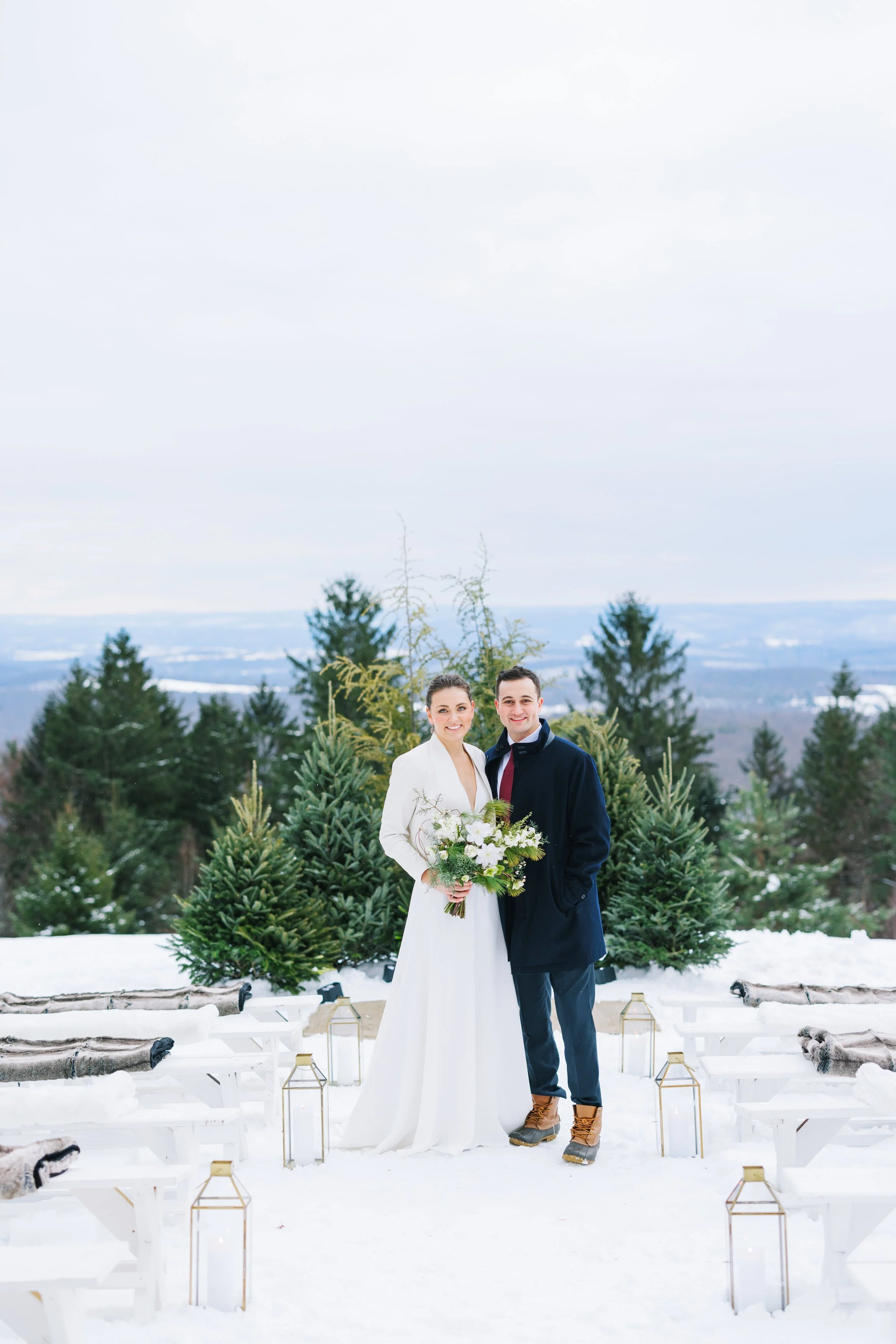 A newlywed couple standing outdoors on snow in winter, with evergreen trees and snowy landscape in background. The bride holds a bouquet, and they are dressed warmly for cold weather.