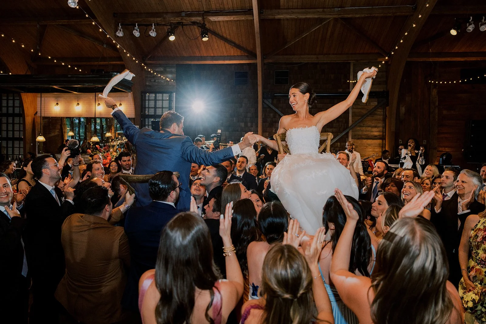 Wedding celebration with bride and groom on the dance floor, bride sitting on a chair with arms raised, surrounded by guests clapping, cheering, and taking photos in a rustic indoor venue with wooden ceiling and string lights.