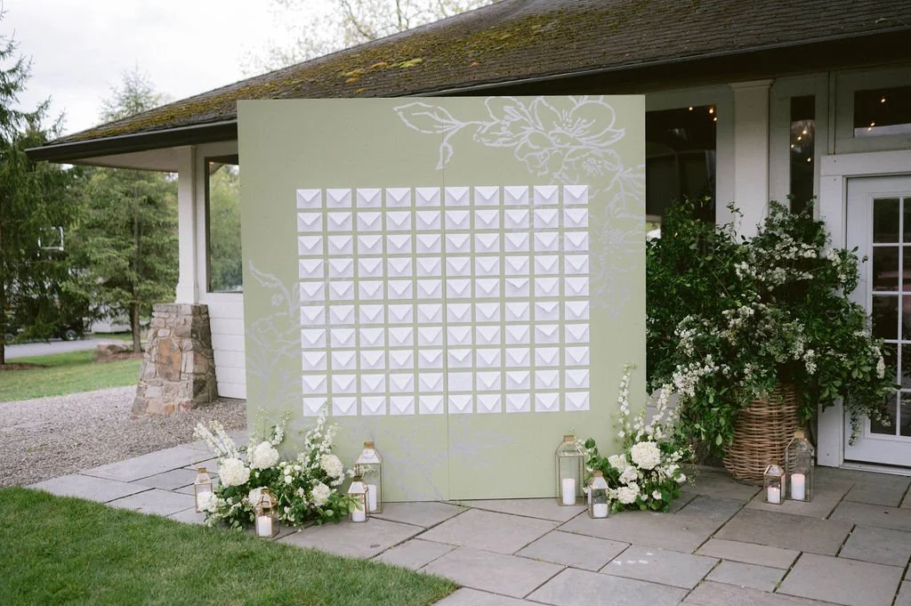A large green backdrop with white 3D geometric shapes, floral illustrations, and surrounded by floral arrangements and lantern candles outside a house.