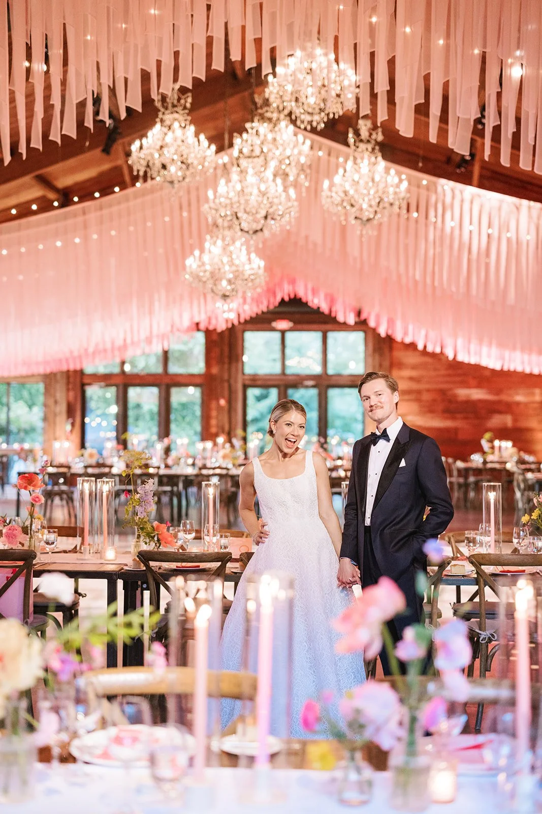 A bride and groom standing hand in hand in a decorated wedding reception venue with chandeliers, pink lighting, and floral table centerpieces.