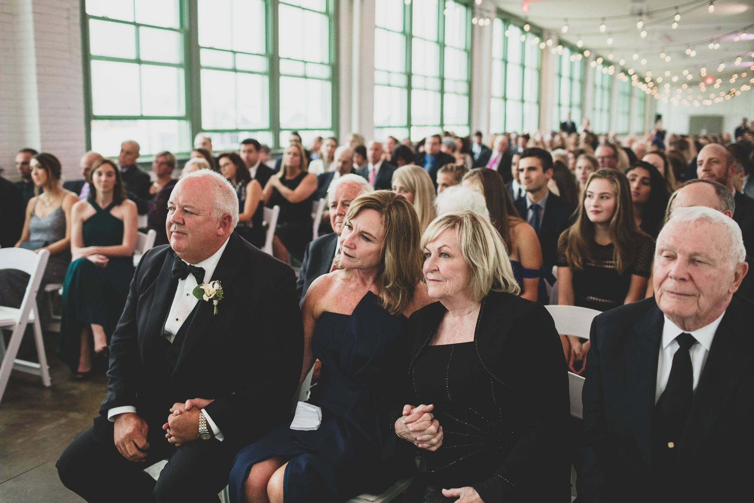 Attendees sitting at a wedding ceremony, with a mix of elderly and young people, dressed in formal attire, inside a decorated venue with large windows and string lights.
