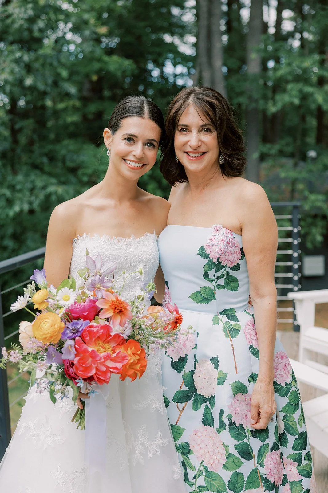 Two women smiling outdoors, one in a white wedding dress holding a colorful bouquet, the other in a strapless floral dress.