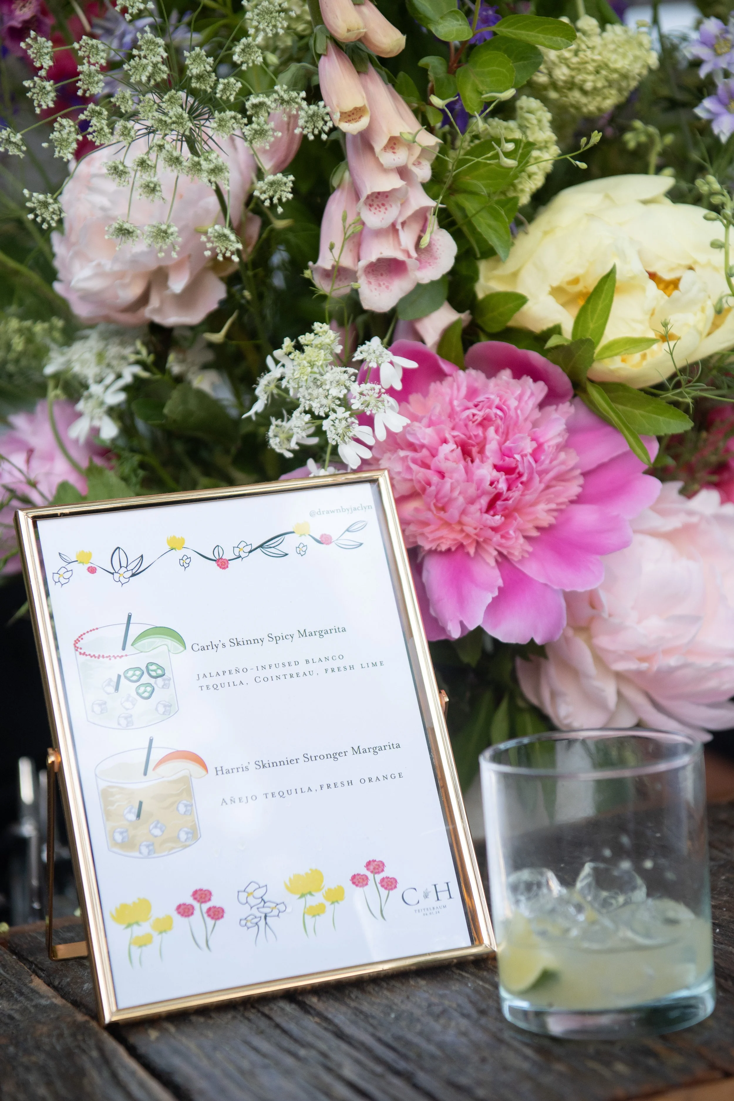 A floral arrangement with pink, yellow, white, and purple flowers, alongside a framed menu and a glass of margarita with ice and lime.