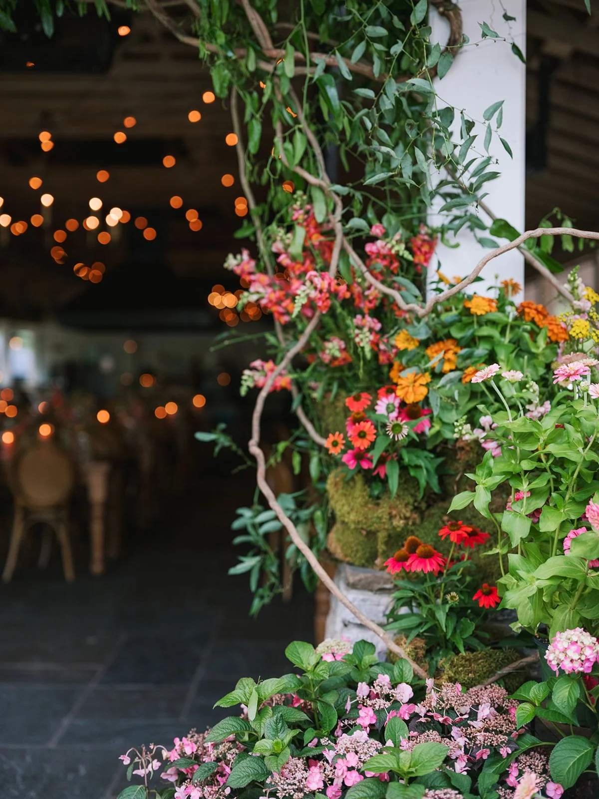 A vibrant floral arrangement with pink, red, orange, and yellow flowers and green foliage at the entrance of a dimly lit venue, with blurred string lights in the background.