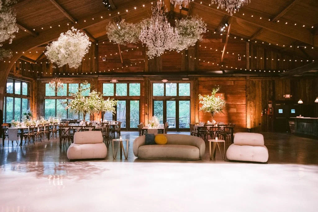 A decorated event hall with wooden walls and ceiling, featuring string lights and floral arrangements, with a cozy seating area in the foreground.