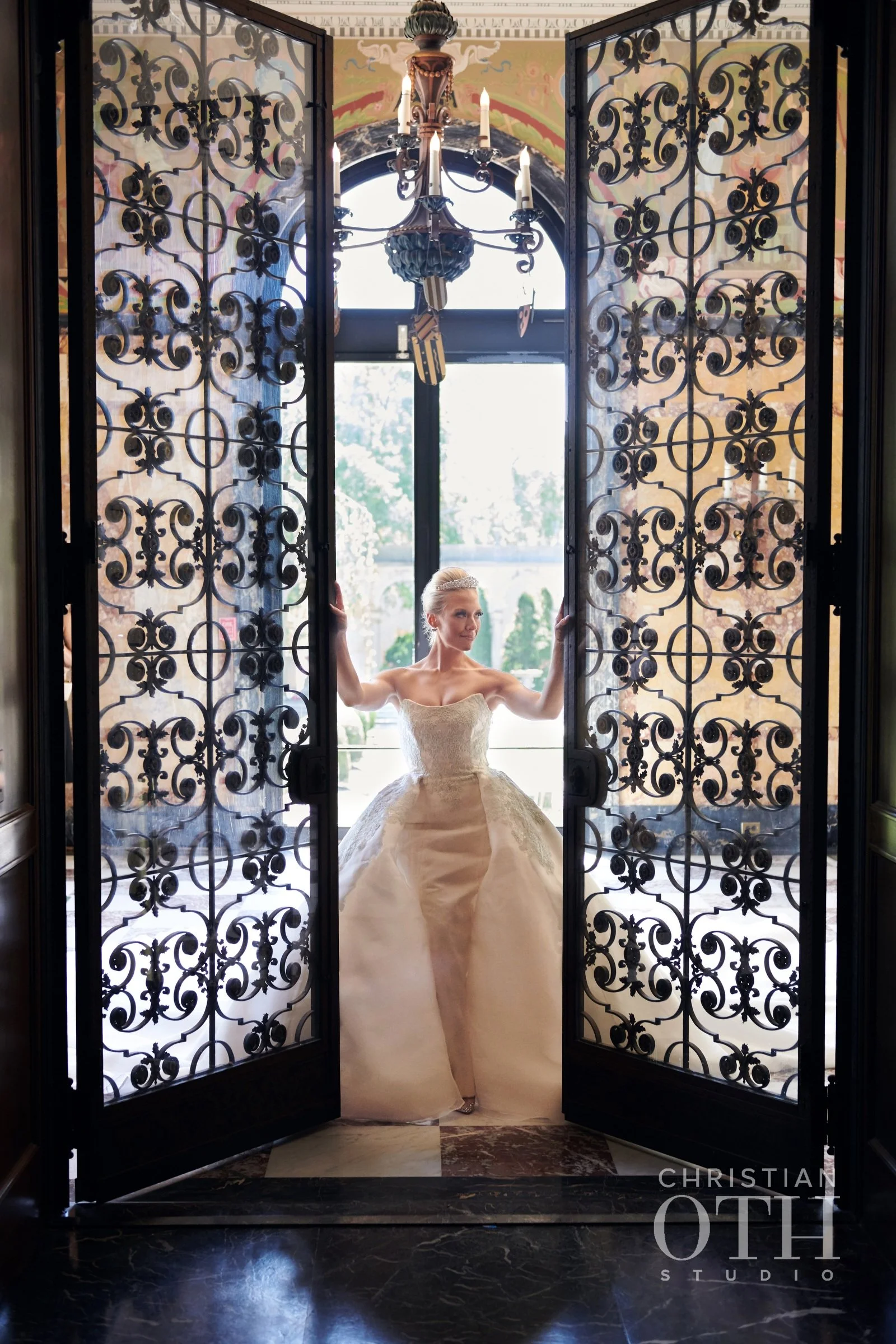 Brunette bride in a strapless wedding dress stands in a doorway with ornate wrought-iron gates, holding the gate open with both hands, in a well-lit room with vintage chandelier and floral decor.