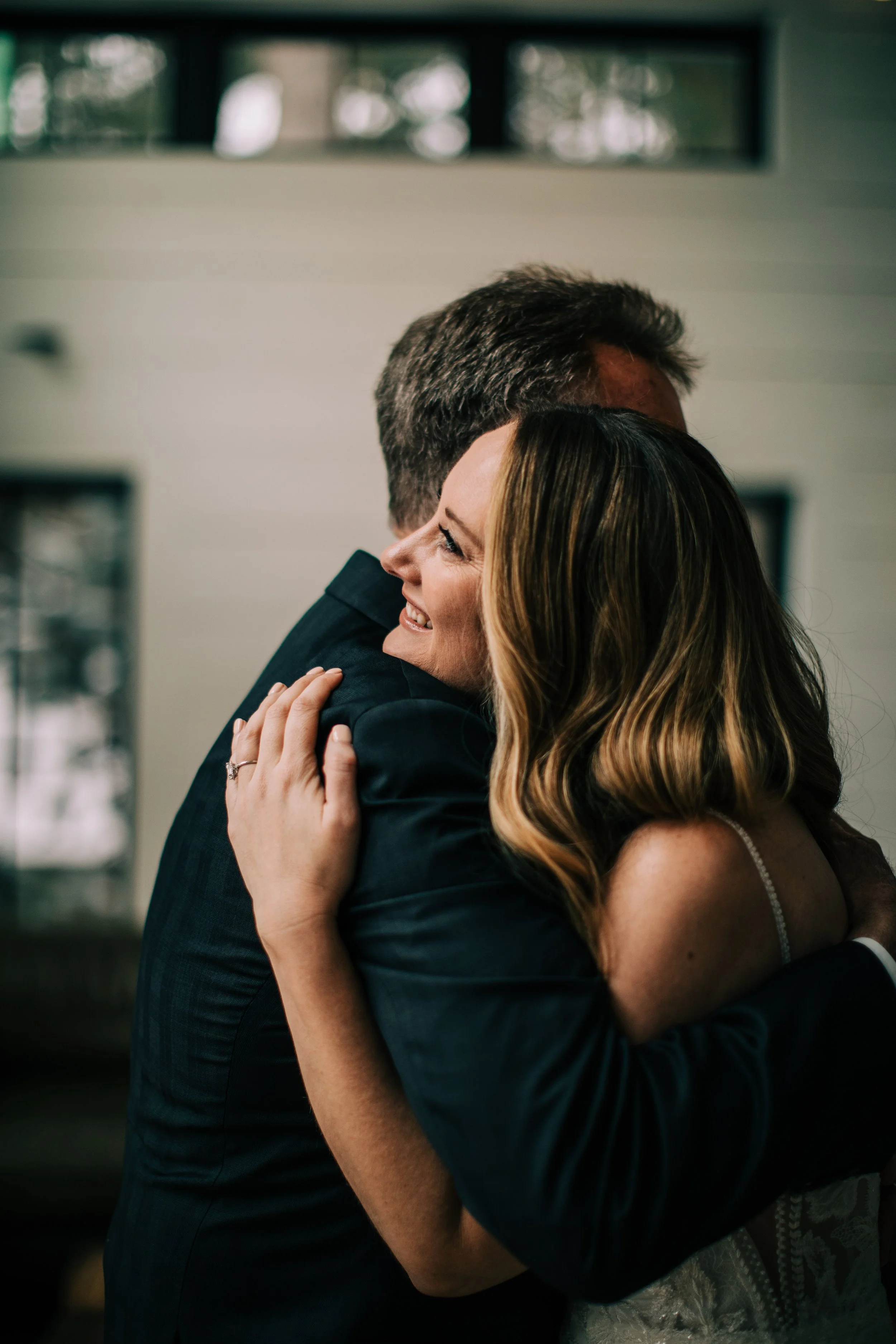 A couple embracing and smiling at each other indoors, with the woman wearing a white dress with lace details and the man in a dark suit.
