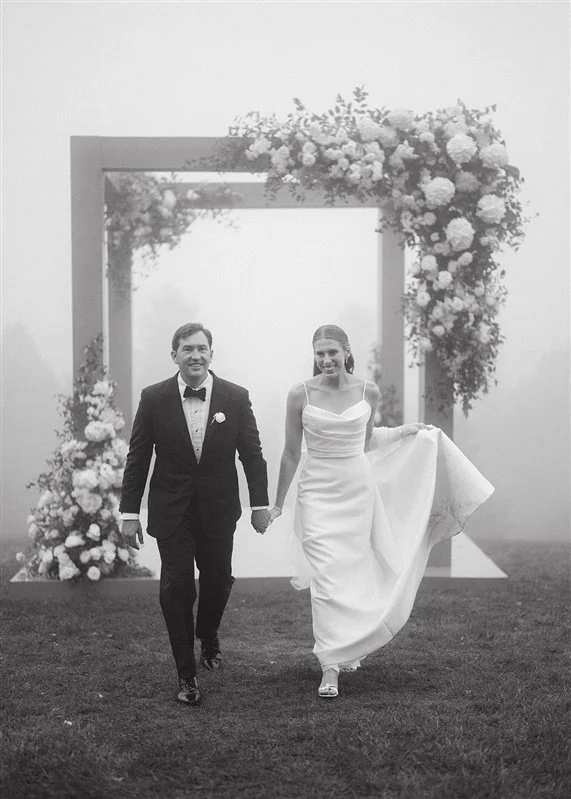 A bride and groom walking hand in hand under a floral wedding arch outdoors on a foggy day.