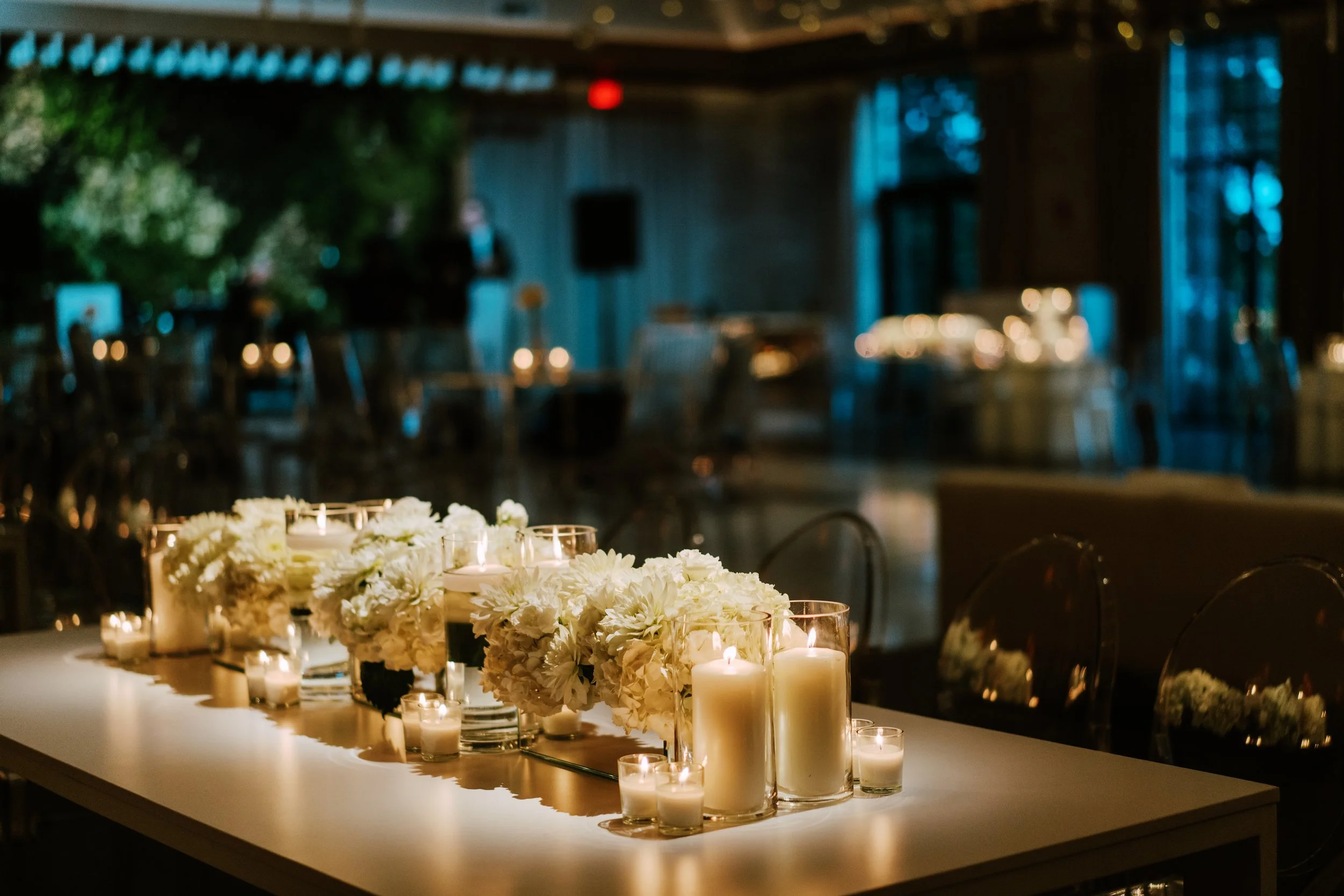 A table decorated with white floral centerpieces and candles in glass holders, set in a dimly lit venue with blurred background and blue lighting.