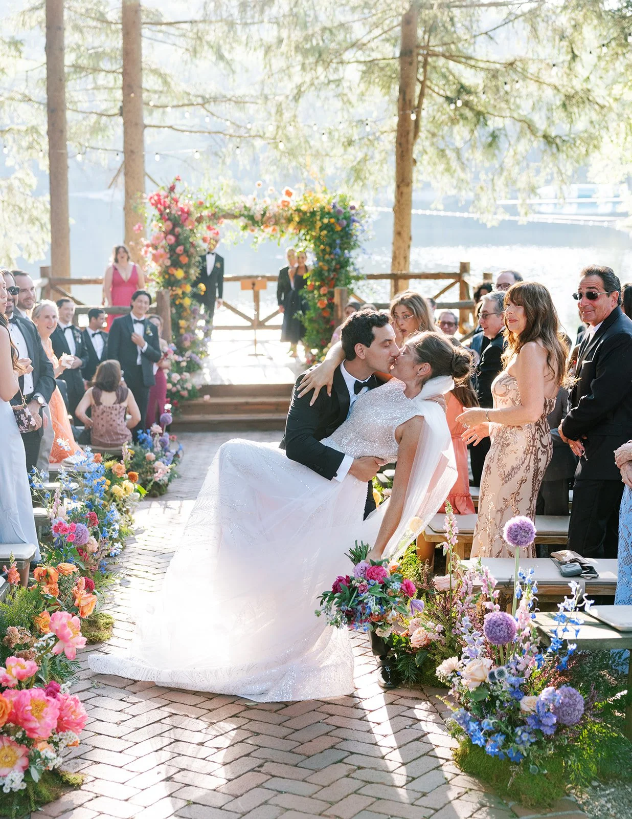 A wedding ceremony with a bride and groom kissing by a lake, surrounded by friends and family, with floral decorations and an arch in the background.