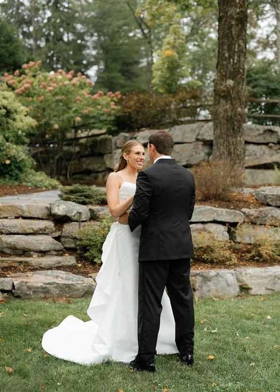 A bride and groom standing outdoors on their wedding day, holding hands and smiling at each other, with trees, rocks, and greenery in the background.