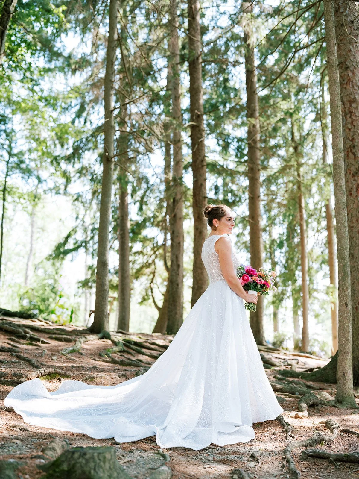 Bride in a white wedding dress holding a bouquet of pink and red flowers standing in a forest with tall trees.