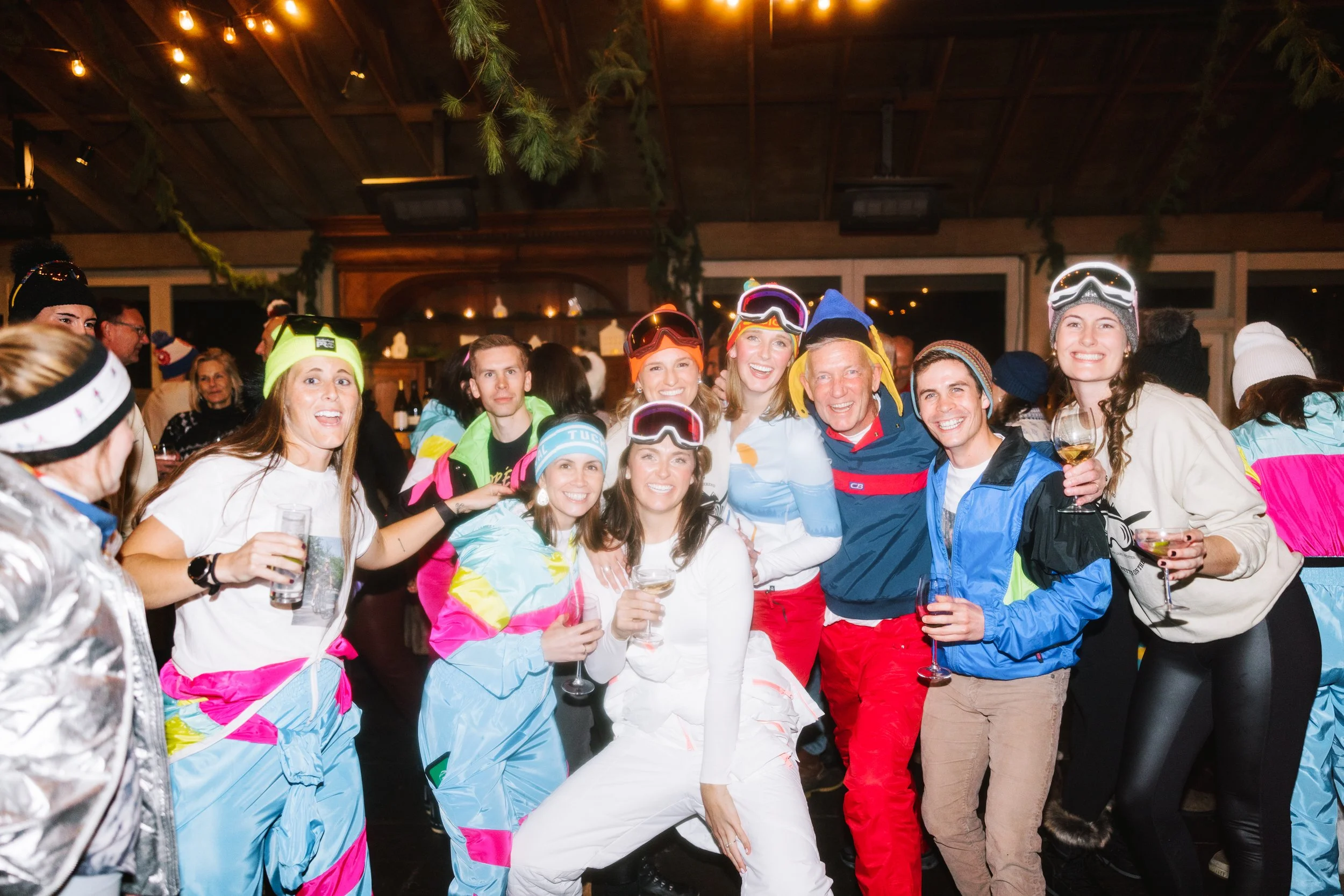 Group of smiling people dressed in colorful ski and winter gear at indoor party, holding drinks, celebrating, with a rustic wooden interior and hanging green decorations in background.