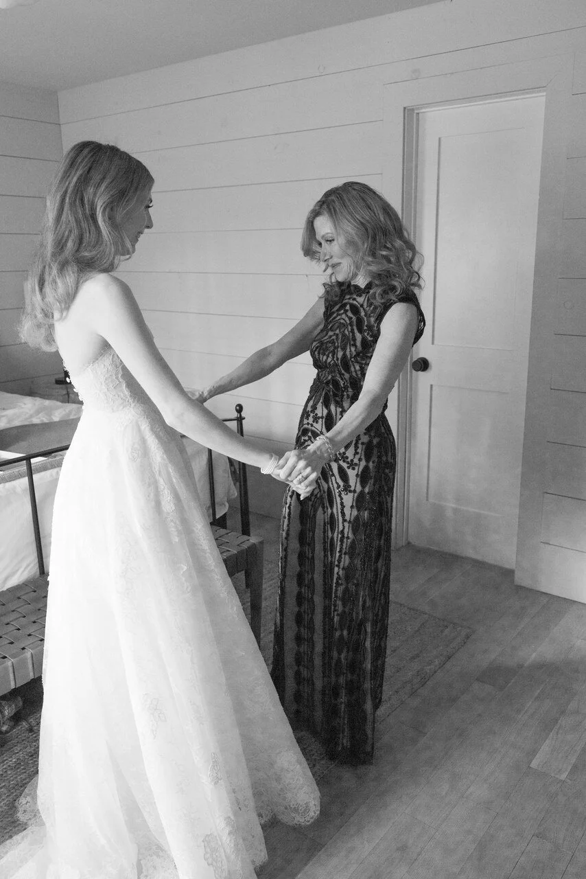 A woman in a black lace dress and a young woman in a white wedding gown holding hands.