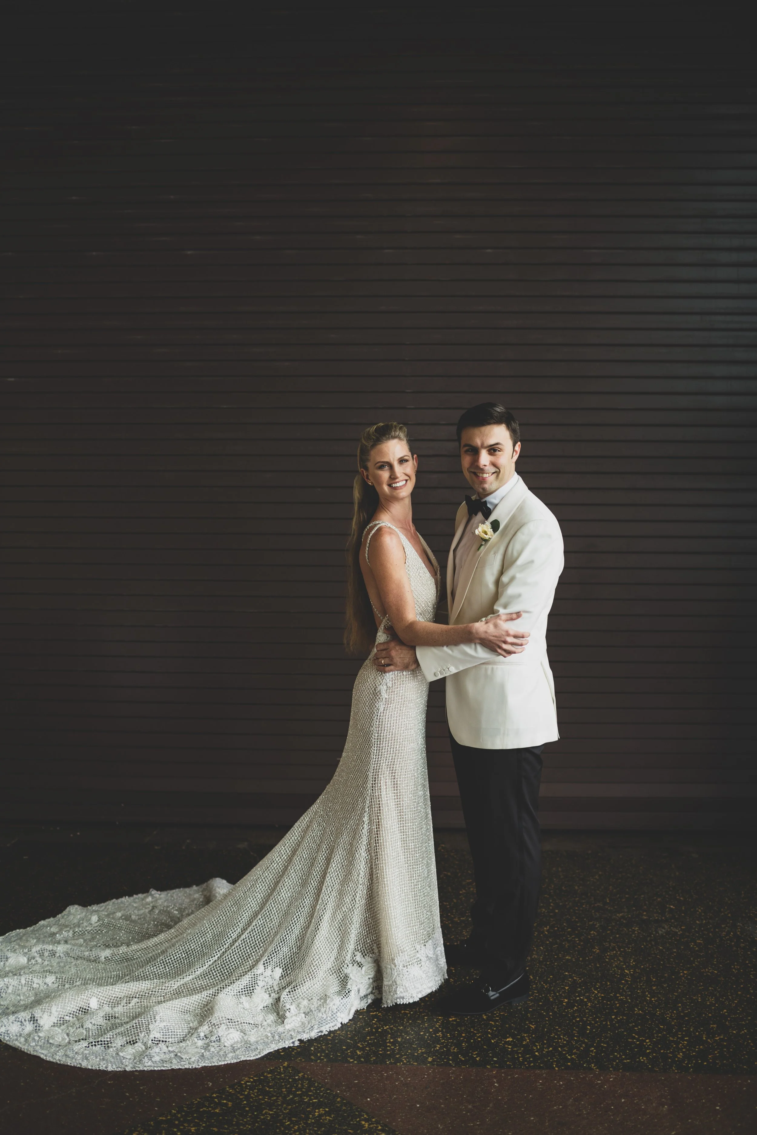 A newlywed couple standing together in wedding attire, smiling, against a dark, horizontal-paneled background.