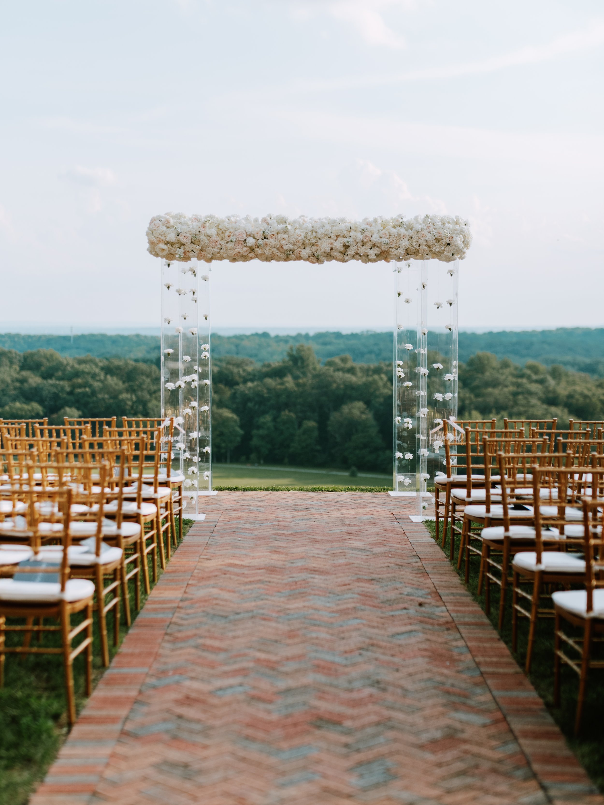 An outdoor wedding ceremony setup with a brick aisle leading to a floral arch, surrounded by gold chairs with white cushions, overlooking a lush green landscape.
