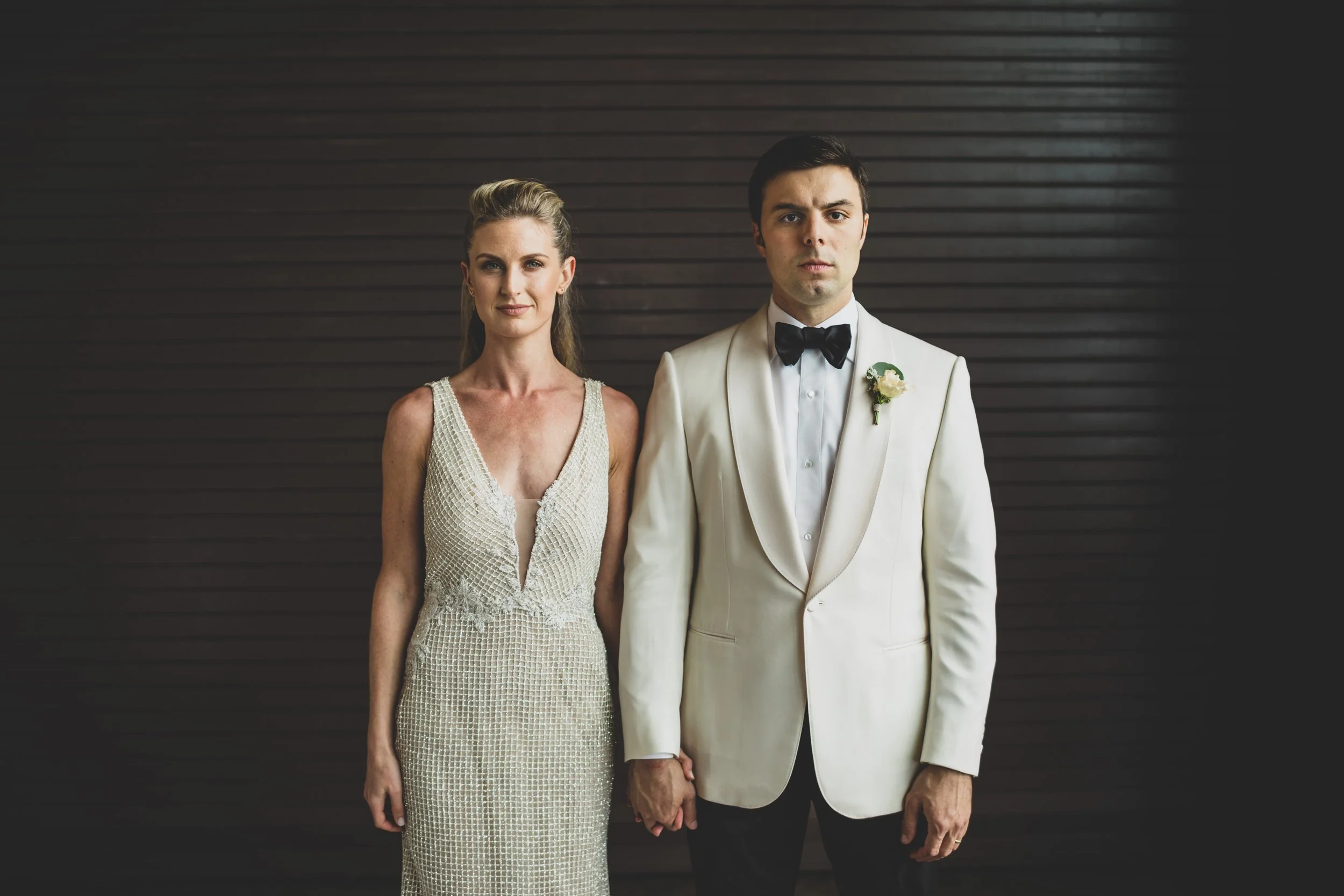 A man and woman dressed in wedding attire standing side by side against a dark wooden background, holding hands.