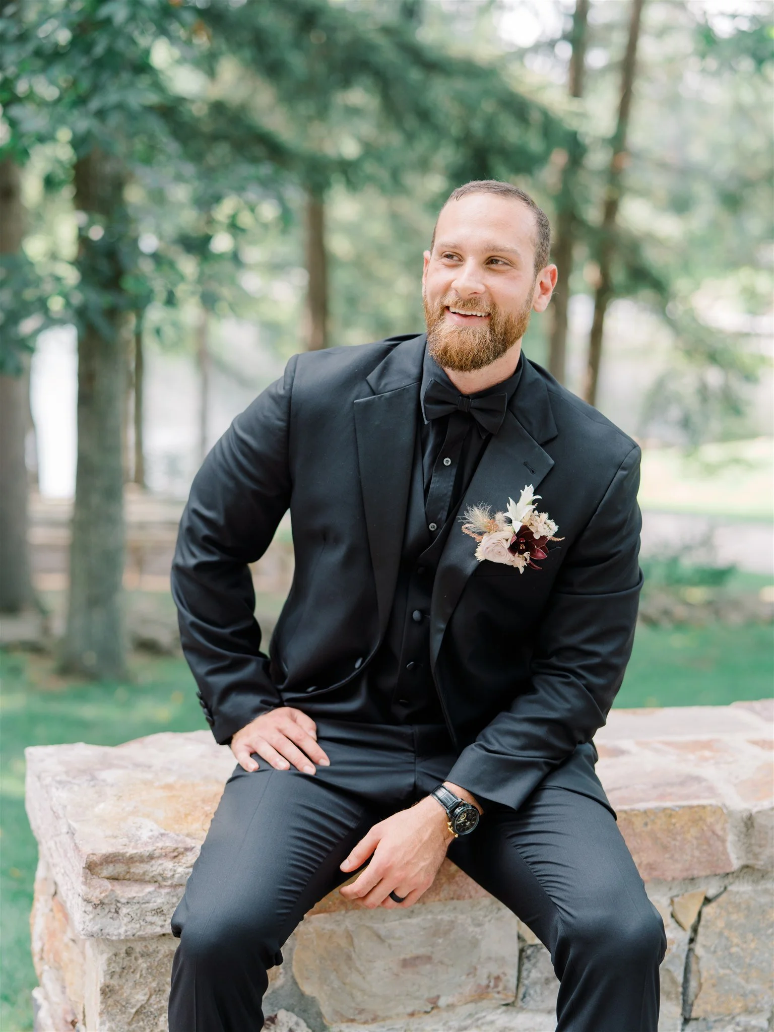 Man in black tuxedo with a boutonniere, sitting outdoors on a stone ledge, smiling, with trees in the background.