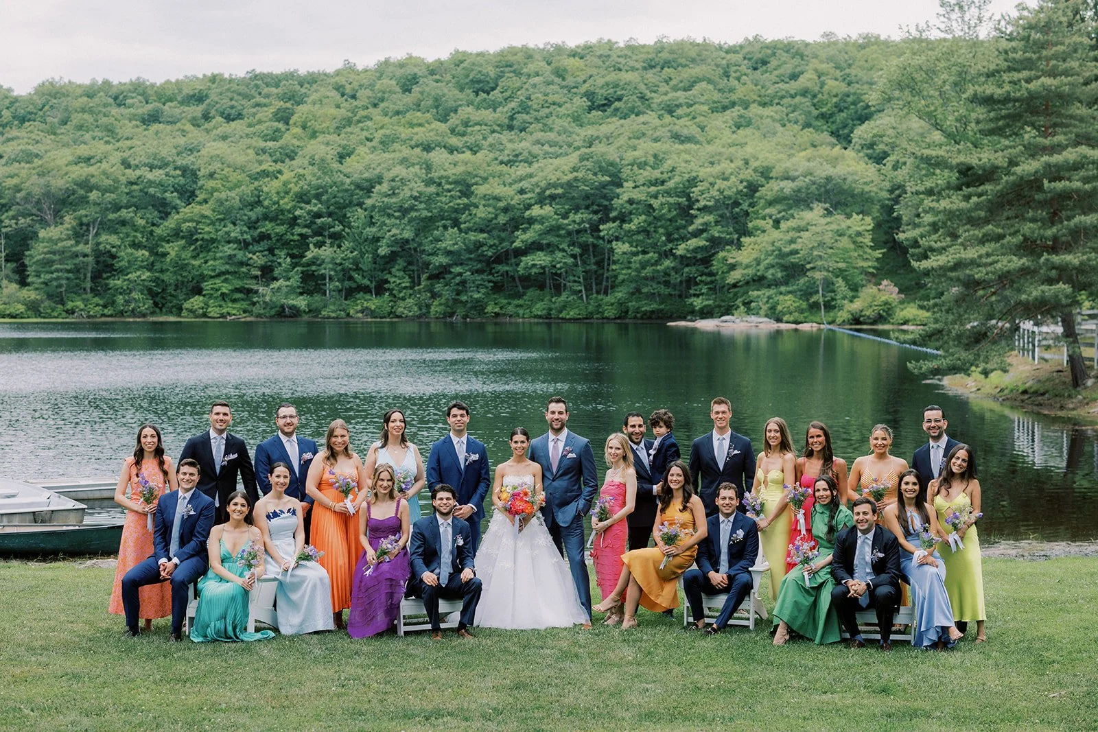 A large wedding party posing outdoors near a lake with lush green trees in the background. The group includes men in suits, women in colorful dresses, and a bride in a white gown holding a bouquet, along with a groom in a blue suit.
