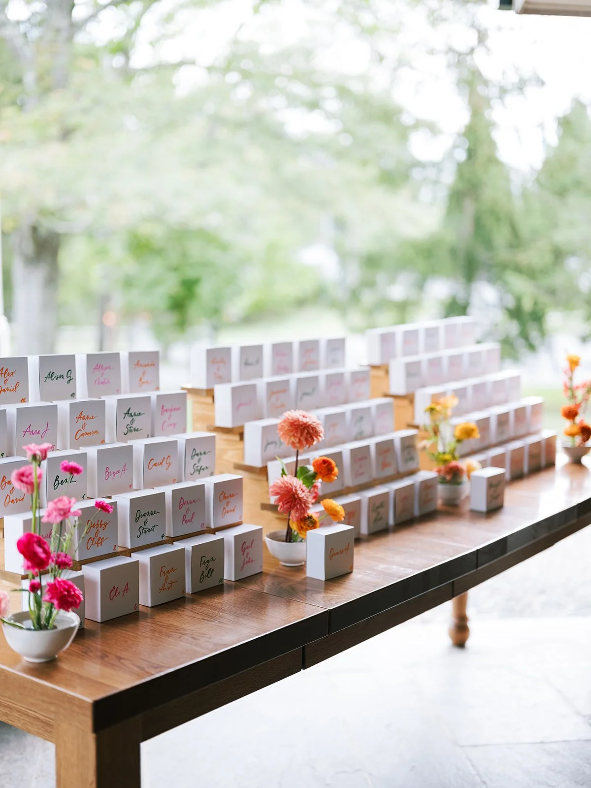 A table displaying place cards and small flower arrangements for an event, with a blurred outdoor scene visible through the window in the background.