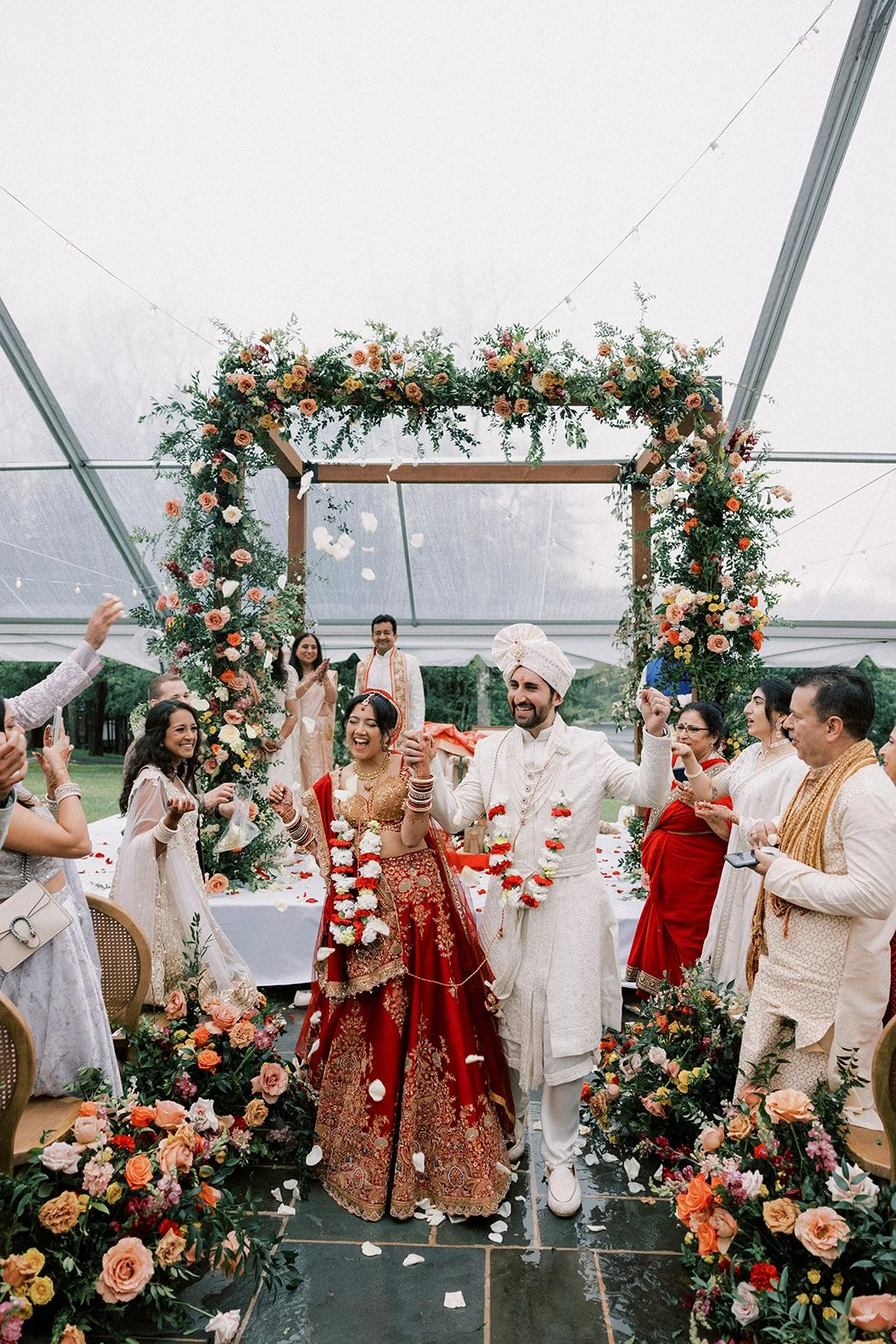 A traditional Hindu wedding ceremony taking place under a decorated floral arch with the bride and groom celebrating. The bride is dressed in a red and gold sari, and the groom is in white traditional attire with a turban. Guests surround them, throw