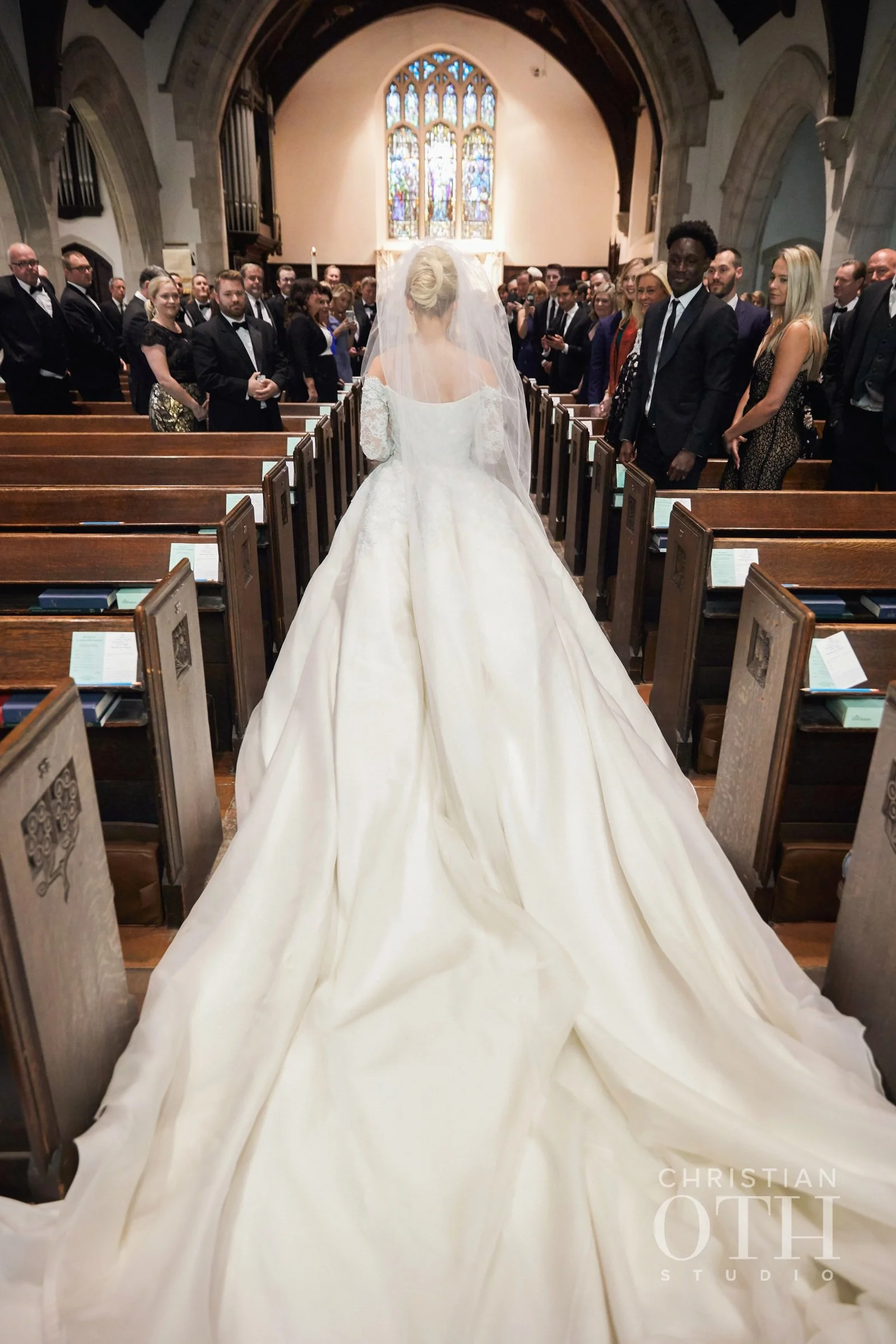 Bride in a white wedding gown walking down the aisle in a church, surrounded by seated guests.