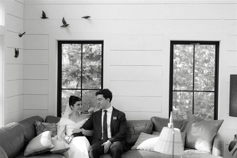 A bride and groom sitting together on a sofa in a cozy room with white walls, two large windows showing trees outside, and decorative birds flying on the wall.