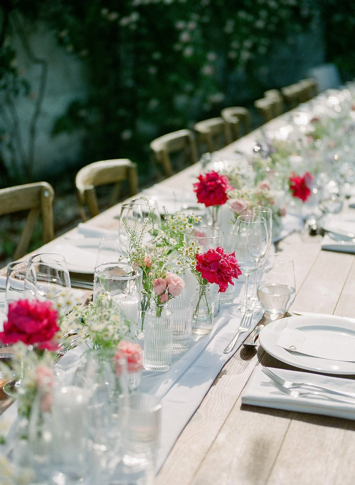 Long outdoor dining table set for a celebration, decorated with pink and white flowers in glass vases, with white plates, silver cutlery, and clear glasses, surrounded by wooden chairs, green foliage in the background, and bright sunlight.