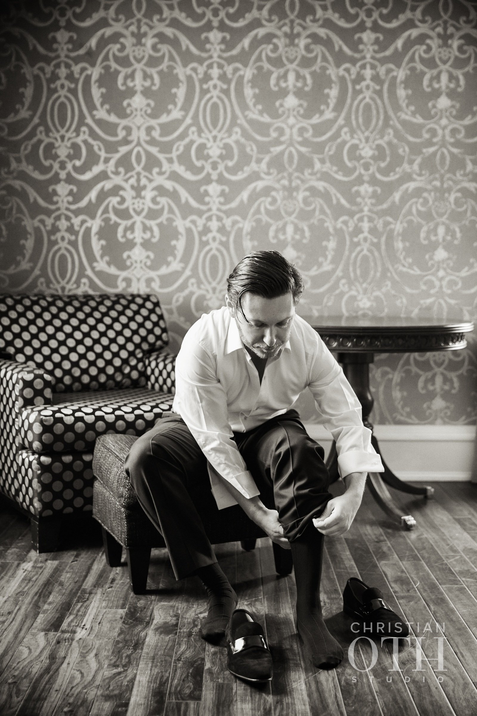 A man sitting on a chair, putting on black dress shoes, in a room with patterned wallpaper, a patterned sofa, and a round table in the background, in black and white.