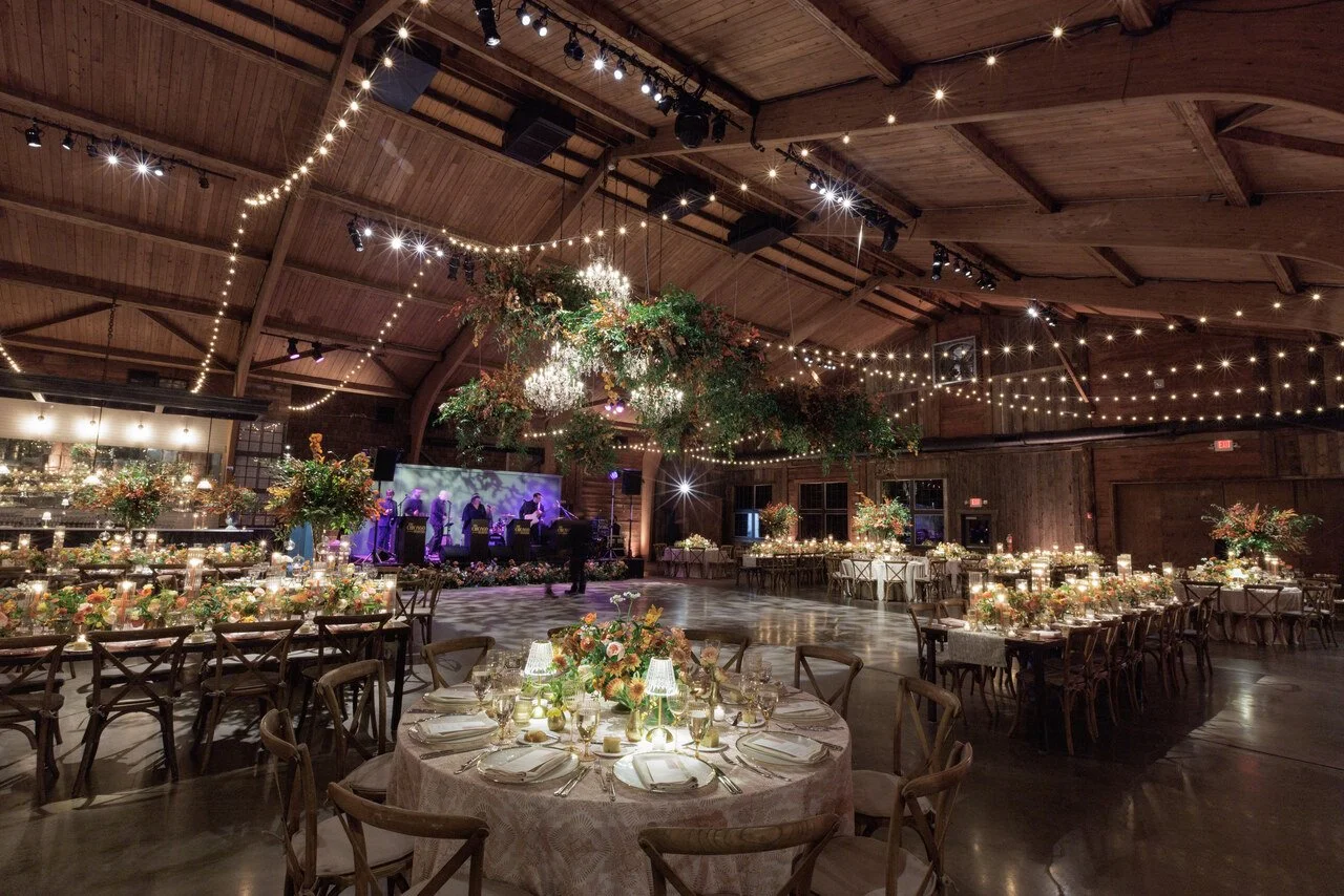Elegant indoor wedding reception hall decorated with hanging string lights, floral arrangements, and a live band stage with musicians. Round tables with floral centerpieces, candles, and place settings are arranged for guests.