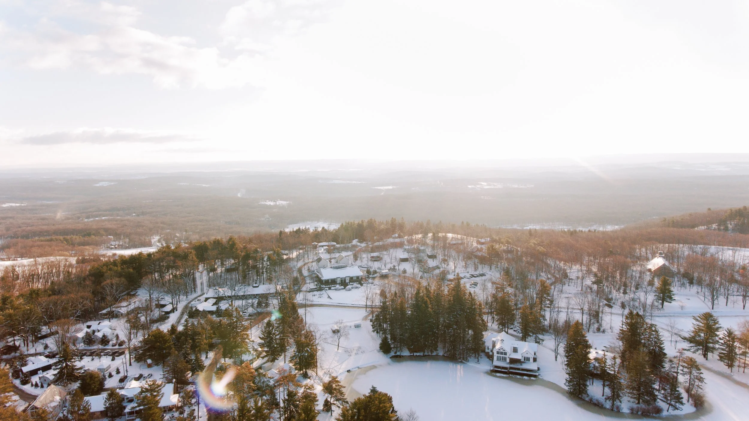 A snowy landscape with houses, trees, and a frozen pond, set in a rural area with rolling hills in the background and a bright sky.