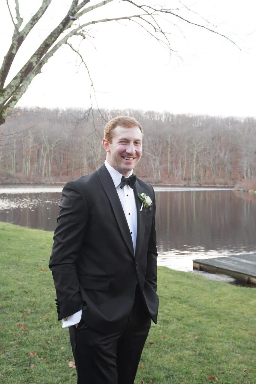 A man in a black tuxedo with a white shirt and black bow tie, standing outdoors near a body of water, smiling at the camera, with a background of leafless trees and overcast sky.
