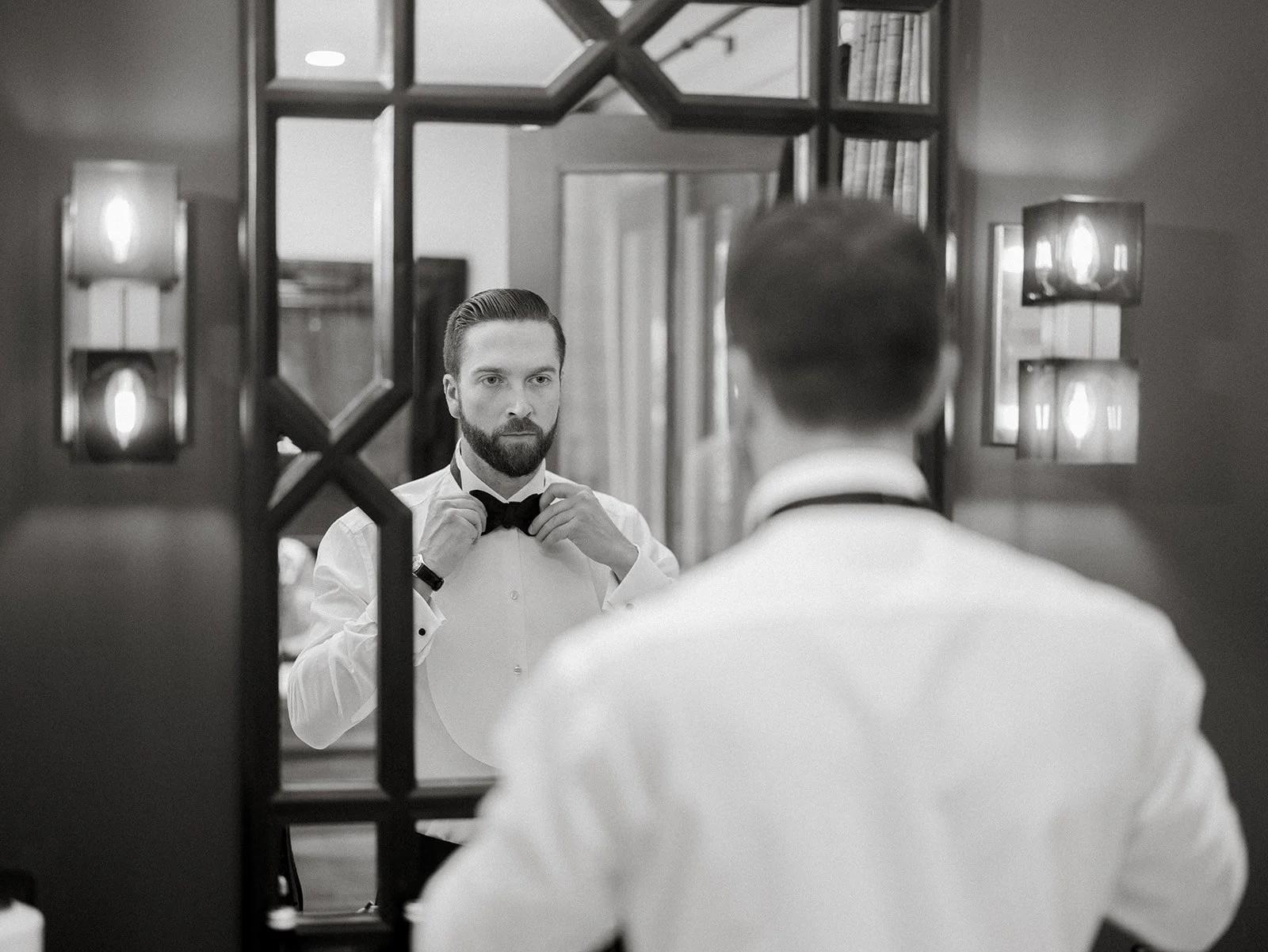 A man with a beard in a tuxedo is adjusting his bowtie in front of a mirror, seen from behind and a reflection in black and white.