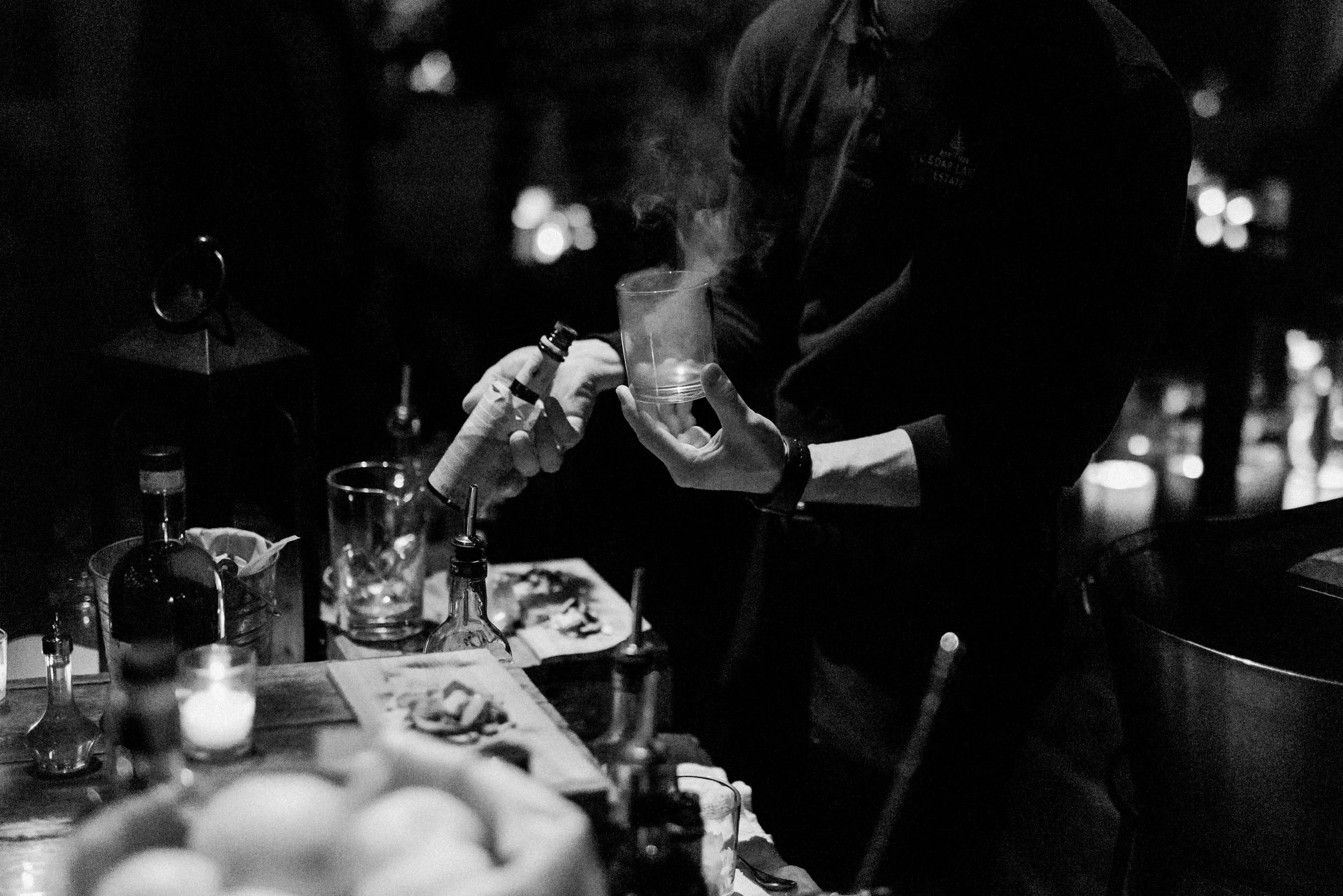 A person handing a steaming glass to another person at a bar in a low-light setting, surrounded by bottles, glasses, and bar snacks.