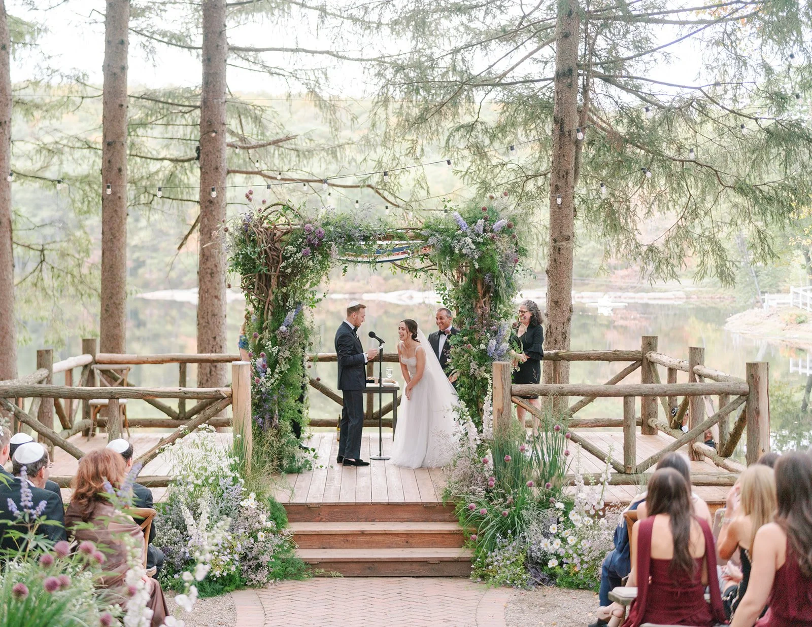 Outdoor wedding ceremony with a bride and groom exchanging vows on a wooden platform decorated with flowers and green foliage, surrounded by tall trees and guests seated on both sides, overlooking a lake.
