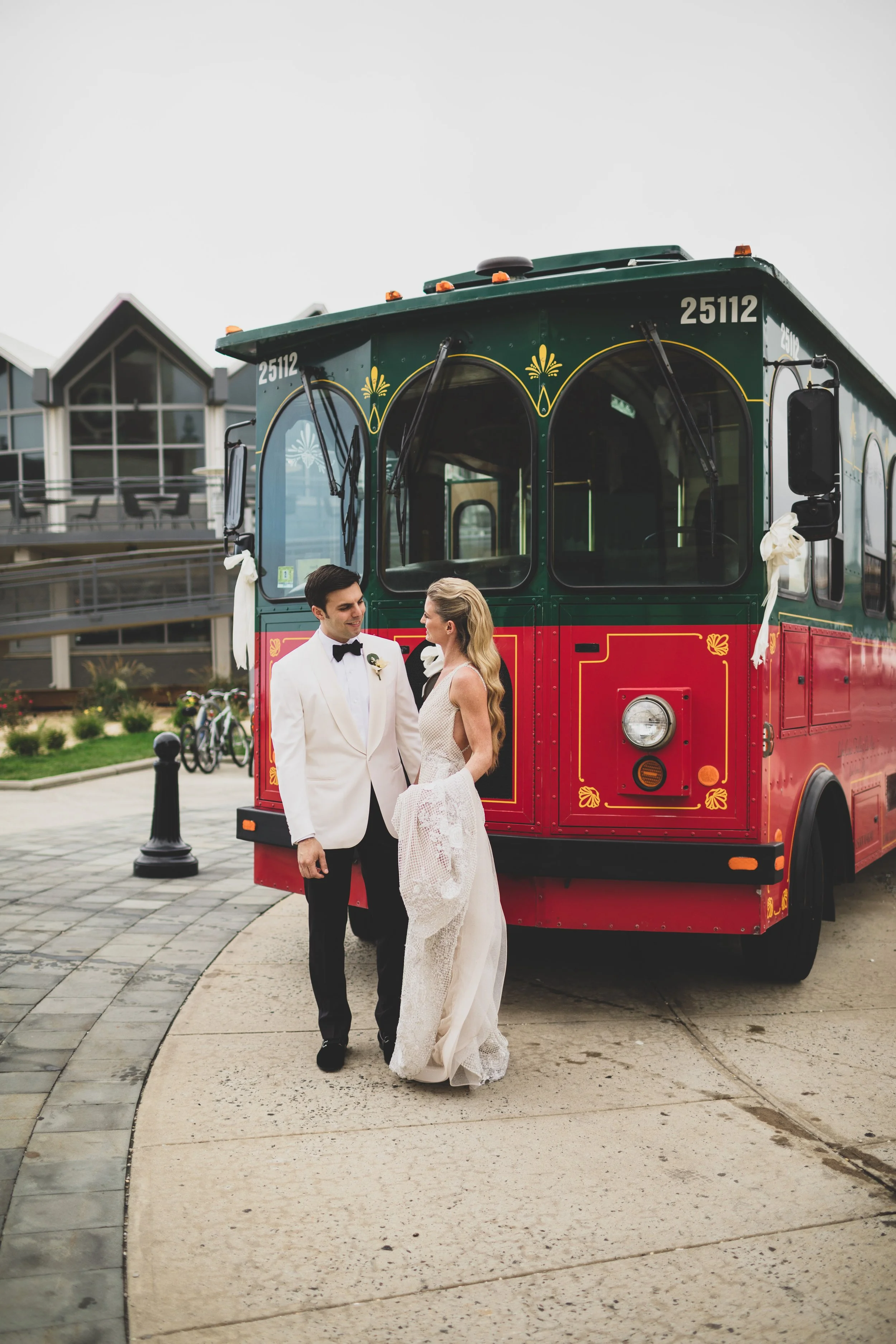 Bride and groom in wedding attire standing in front of a colorful trolley bus, smiling at each other.