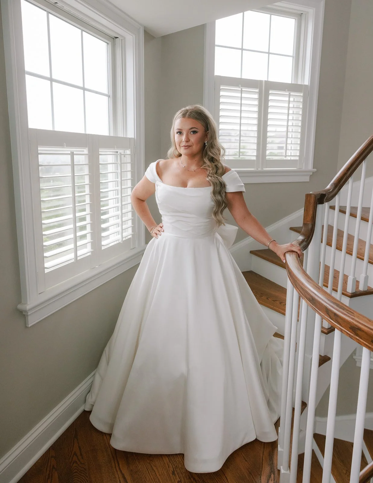 A woman in a white wedding dress standing on a staircase indoors, with large windows and white shutters in the background.